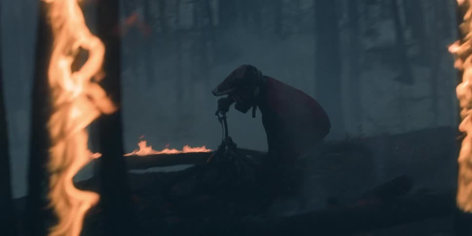 A person wearing a helmet rides a mountain bike through a smokey burning forest with trees in flames