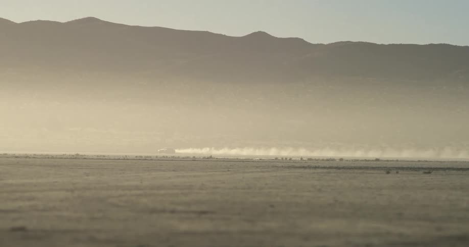 Sports car leaving a dust trail while driving on the desert close to the mountains at sunset