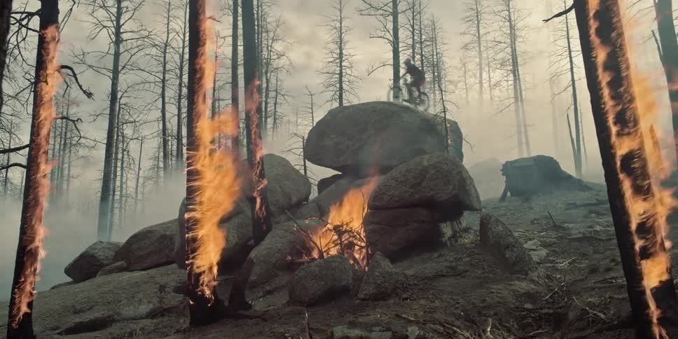 Mountain biker jumps on to rock and off again through burning trees.