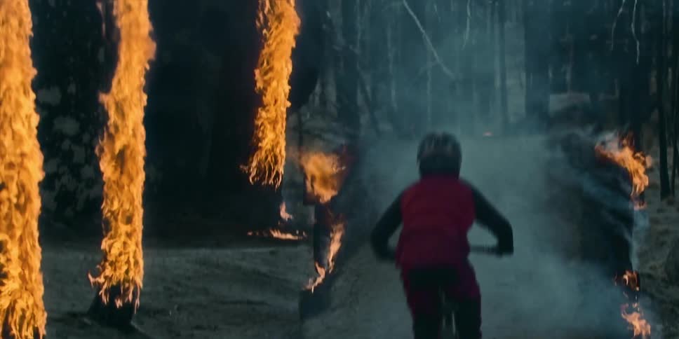A person wearing a helmet rides a mountain bike over a ramp and flies through the air in aburning forest with trees in flames