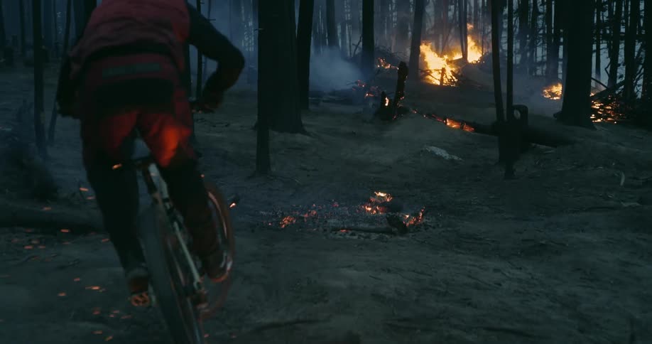 A person wearing a helmet rides a mountain bike through a smokey burning forest with trees in flames