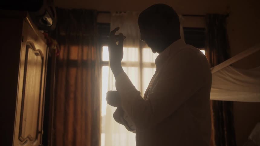 A man buttons the sleeve of his white shirt while standing near a window in front of a wooden wardrobe closet
