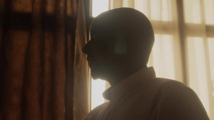 A bald man stands in front of a window with bright sunlight coming through sheer curtains