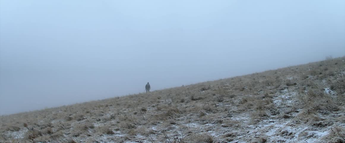 Person walking across dry grassland as snow falls