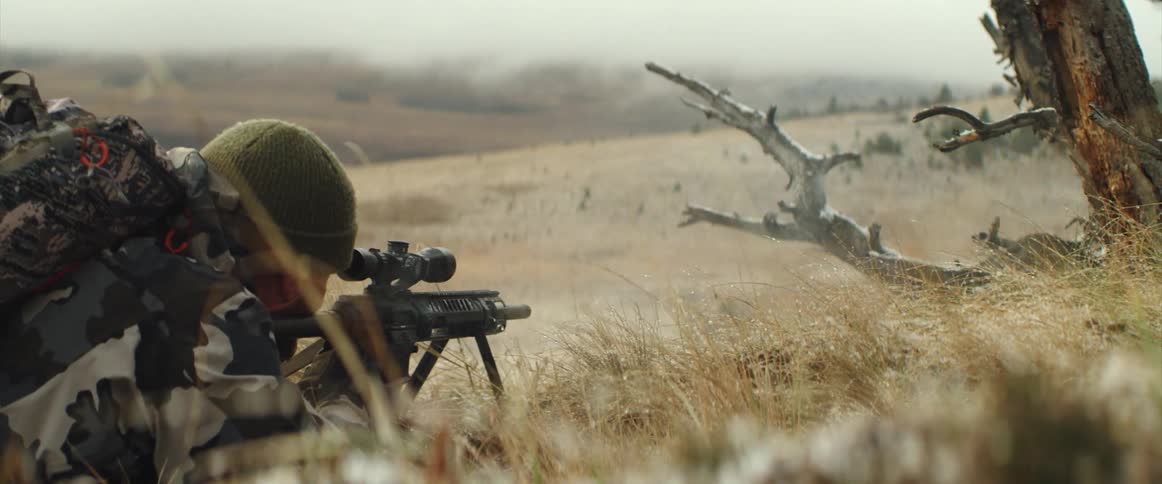 Soldier laying over the dry grass on a field fires an assault rifle