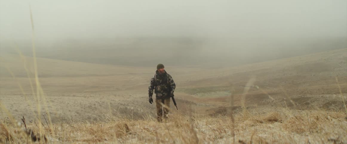 Man wearing camouflage walks across fog covered terrain carrying a rifle