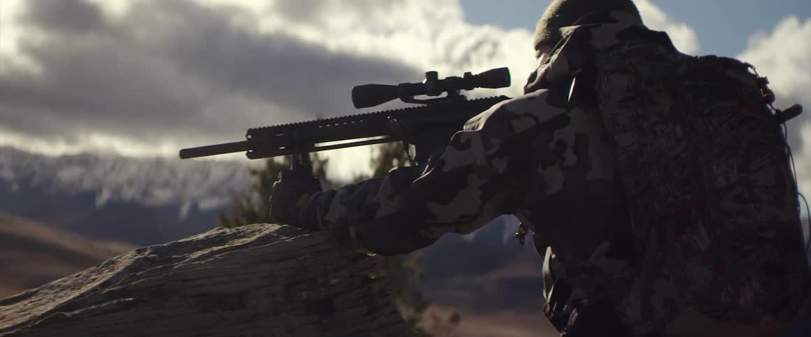 A man with fatigues and a backpack shooting a rifle with a scope with the sky and clouds in the background