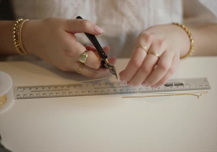 A woman sits at a table in front of a ruler where a gold necklace is lined up and clips it with a pair of clippers