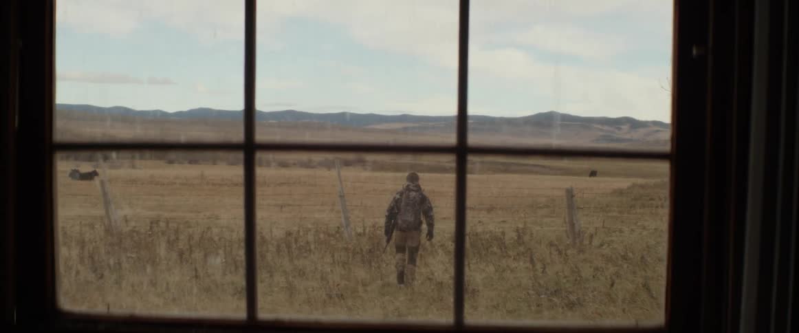 A window with a person with a rifle walking on a grassy plain with a mountain range the sky and clouds in the background