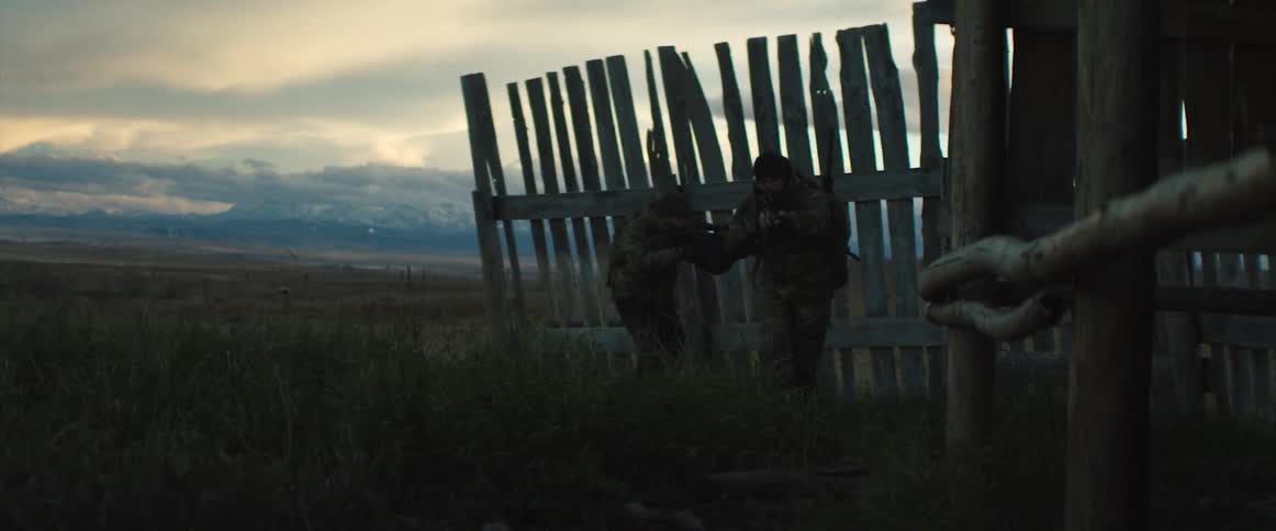 Two soldiers with assault rifles walking near a wooden fence on a field