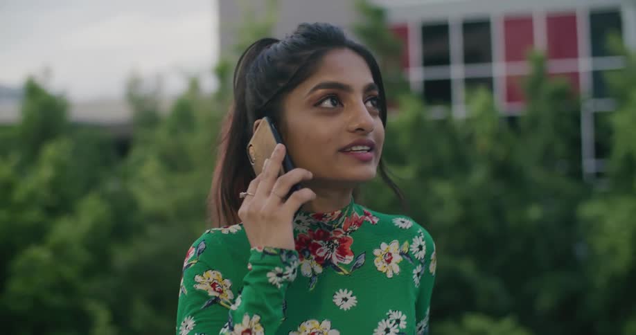 A young woman speaks on a smartphone while standing outdoors outside of a house during the day