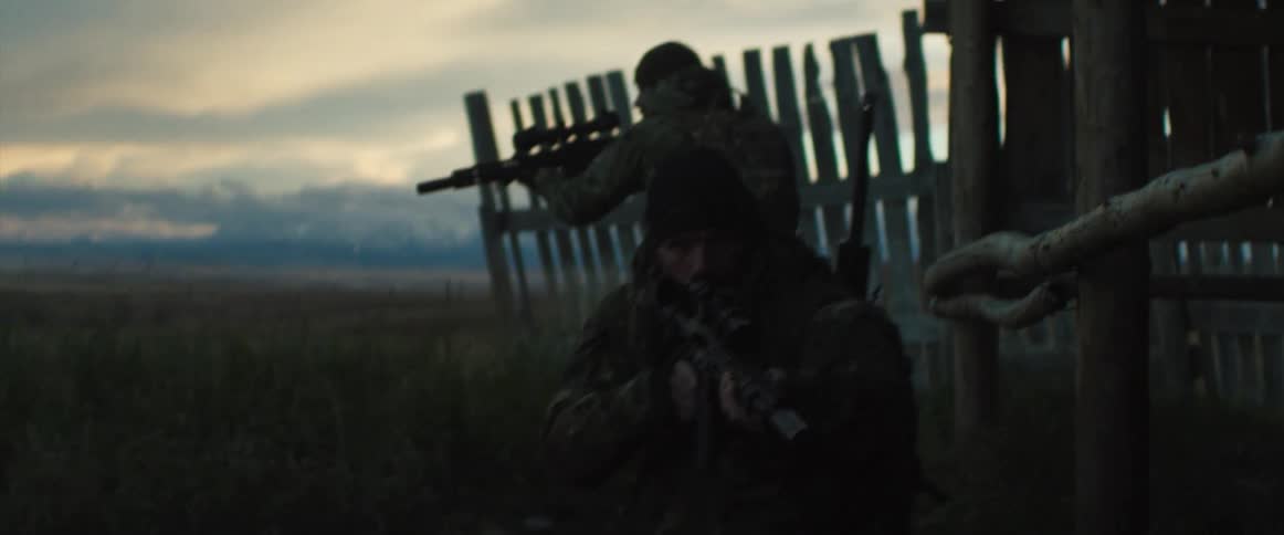Two men with rifles walking on a grassy field next to a fence with the sky and clouds in the background