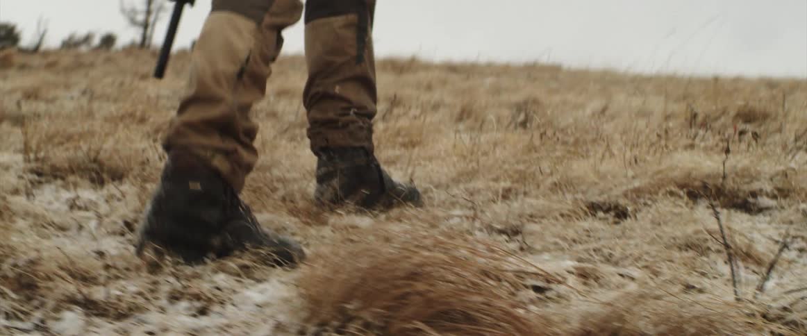 A person with a rifle walking on a grassy plain with trees the sky and clouds in the background