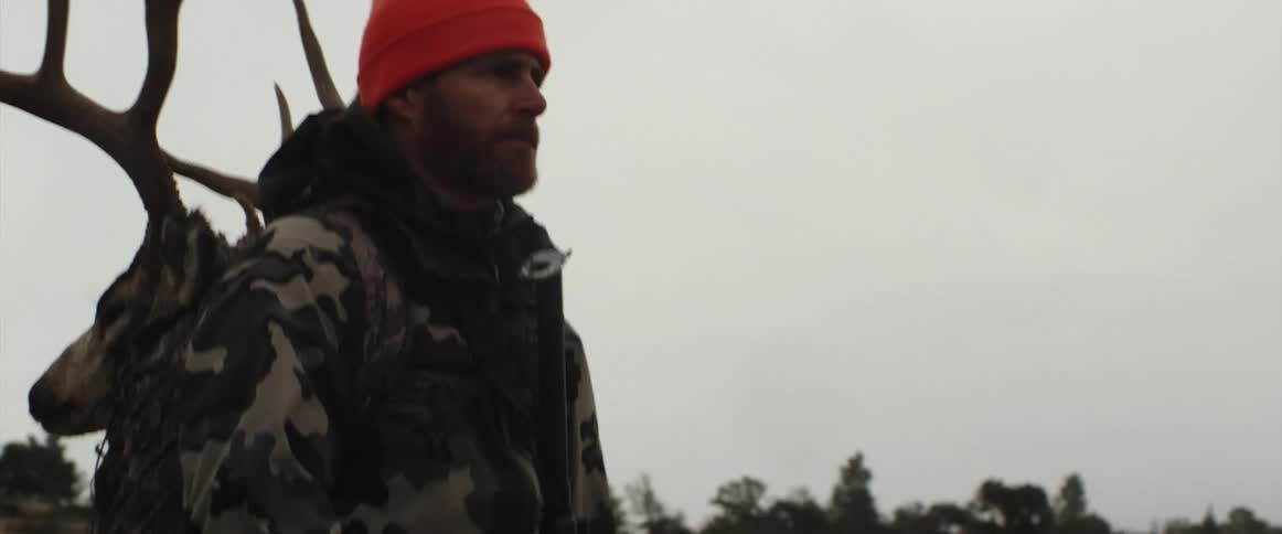 A man in hunting gear walks across a field with a deer head on his back on a cloudy day