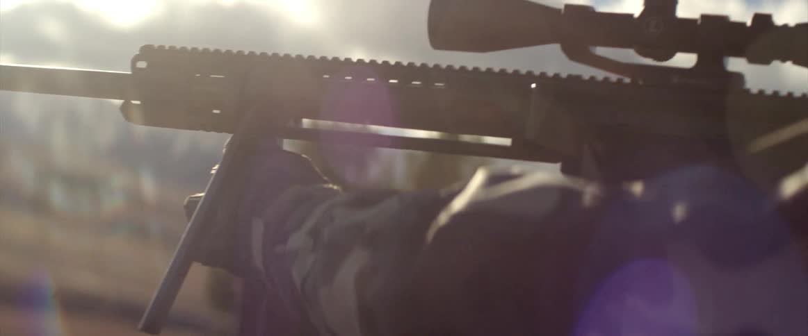Person sets rifle with scope up on tripod on sunny day with mountain slope in distance