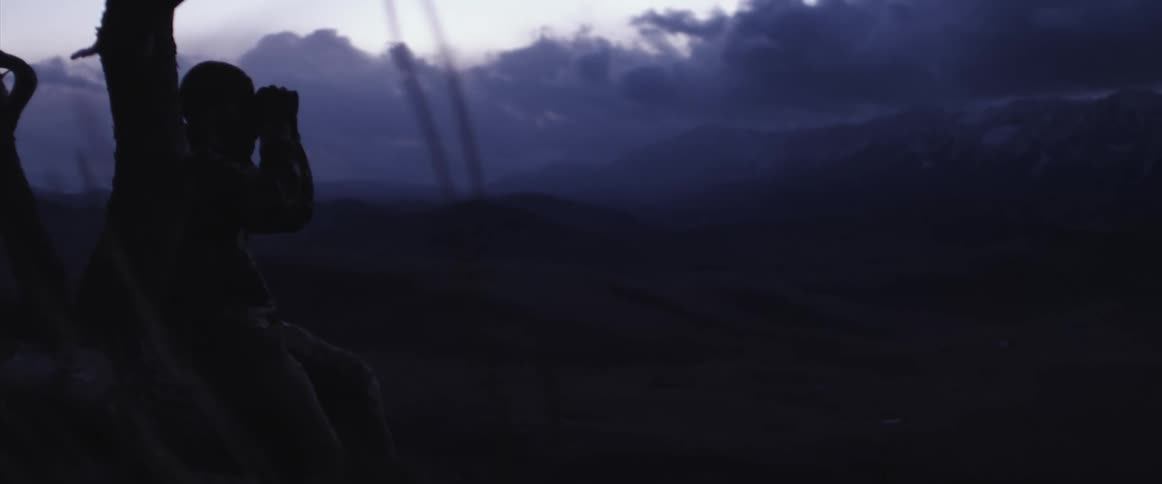 A person uses binoculars on the mountain under dark clouds at sunset