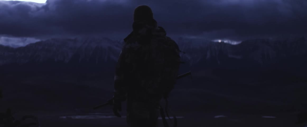 Man holding rifle stands looking toward shadowed mountains on horizon