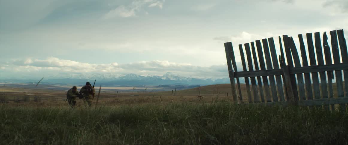 Two men with rifles walking on a grassy field with a fence in the foreground and the sky and clouds in the background