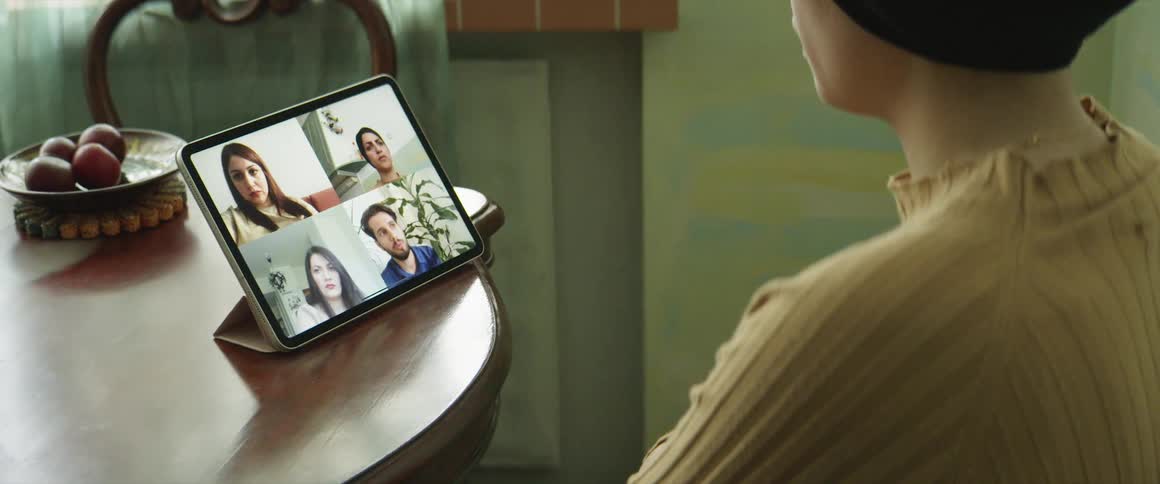 A woman sits at her dining room table while listening to a video call on her tablet with four people