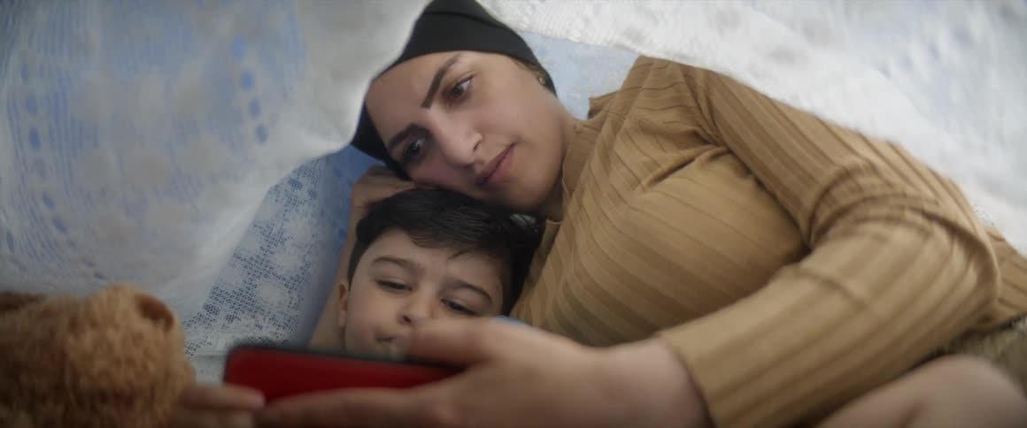 A mother and her son cuddle up under a blanket and watch a screen together