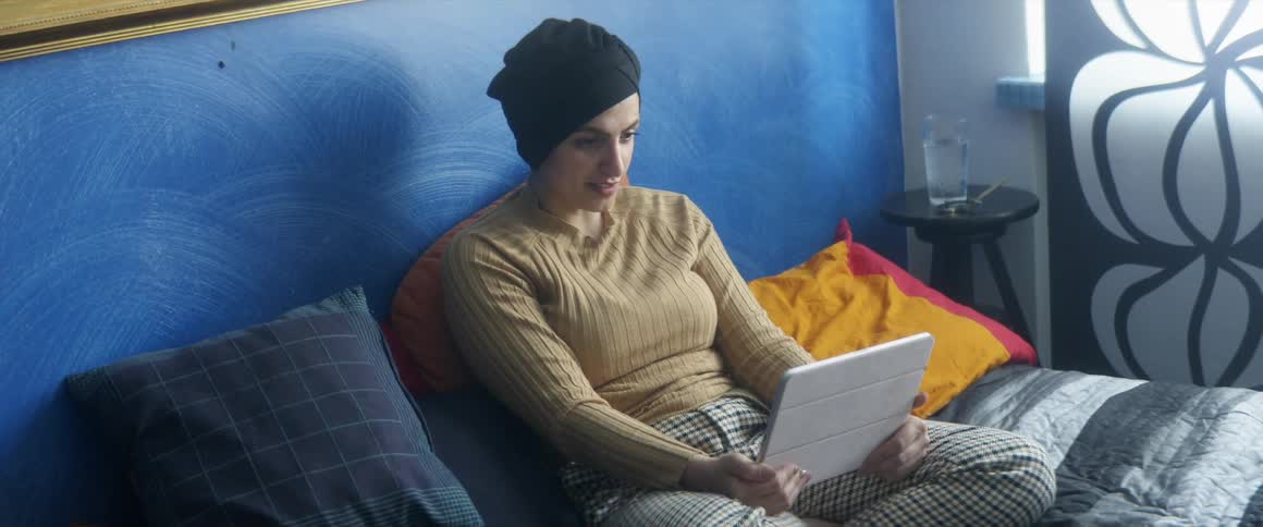 Woman with a black headscarf a brown sweater sits on the bed and uses a tablet in the bedroom