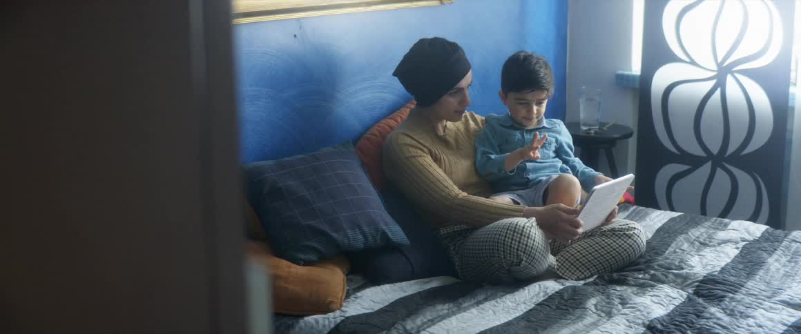 A mother and son sit together on a bed as they look at a tablet in a bedroom during the day