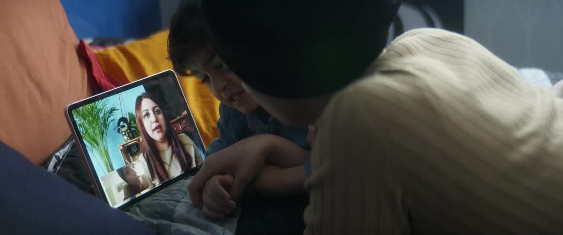 A woman attempts to stop her son from fiddling with her tablet as she video chats with another woman while lying on her bed