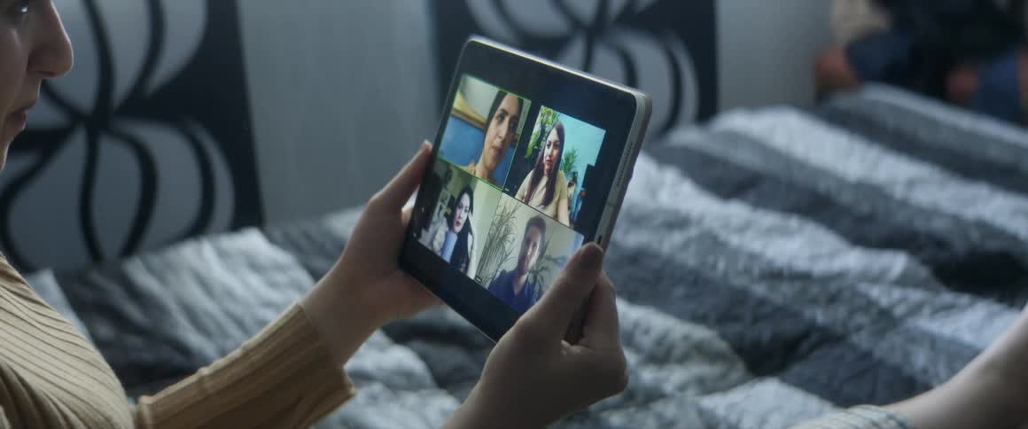 A woman uses her tablet to video chat with other people while sitting on her bed during the day