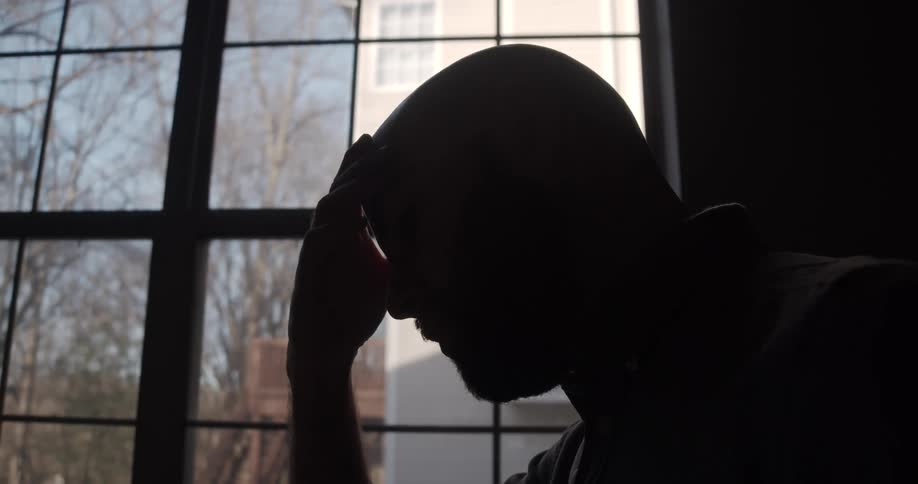 A depressed man strokes his forehead while sitting near a window in a dark room