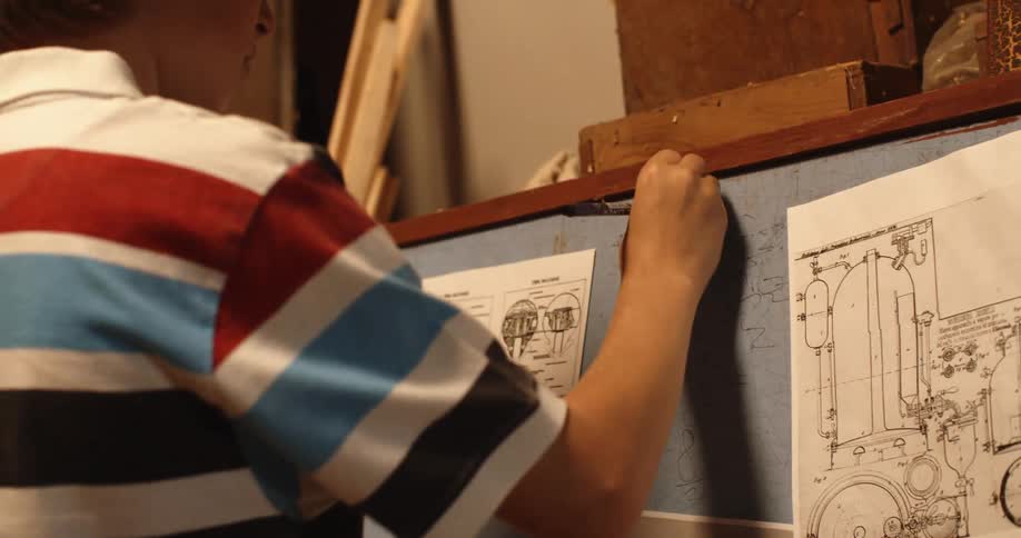 Young blonde boy writing on a blackboard filled with blueprints