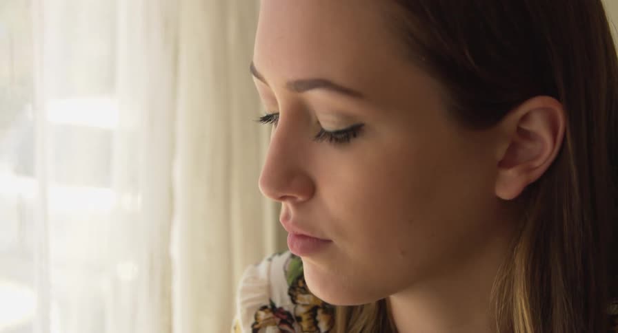 A young woman rests next to a curtained window indoors during the day