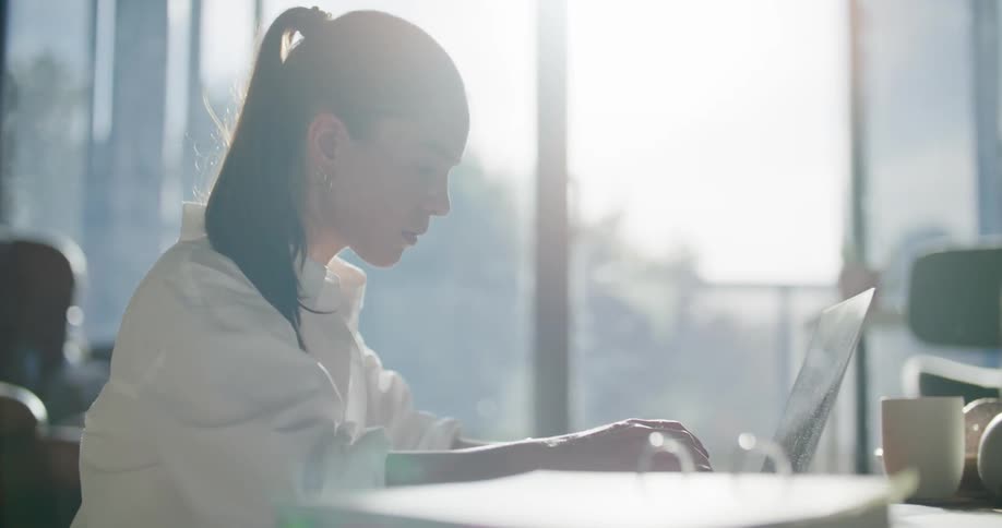 Woman types on laptop consulting papers in bright office