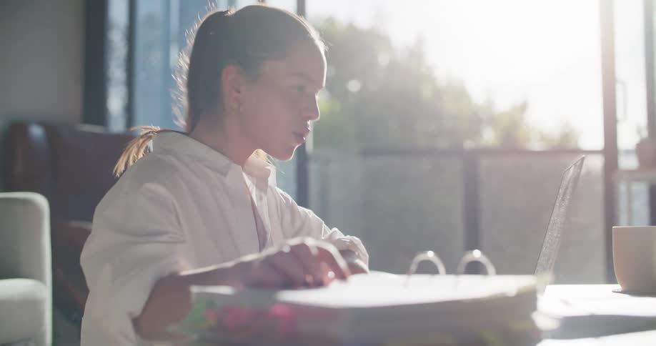 A woman sitting at a table video chatting on a laptop with a couch and a balcony in the background