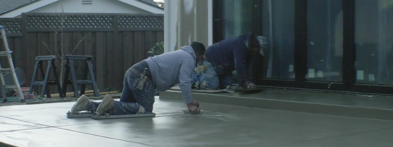 Two men smoothing cement with a ladder and a fence in the background