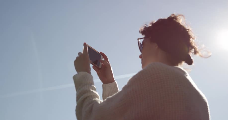 A woman with brown curly hair takes video on her phone while standing outside on a sunny clear day