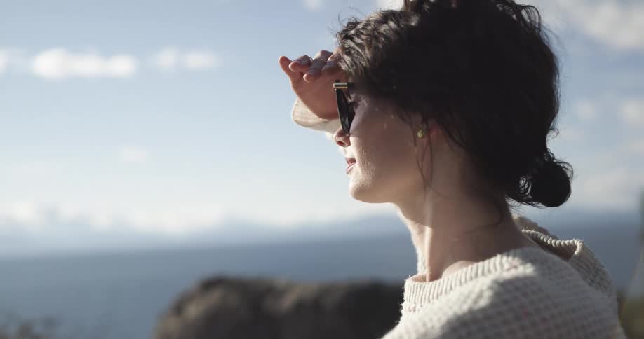 Side view of woman with sunglasses and white sweater looking around on a cliff edge