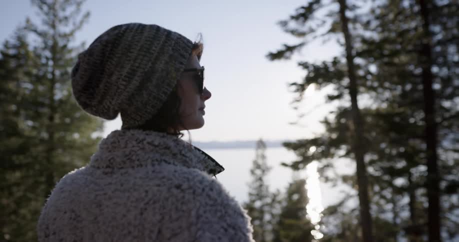 Woman gazes past conifer trees toward water and mountains on opposite shore