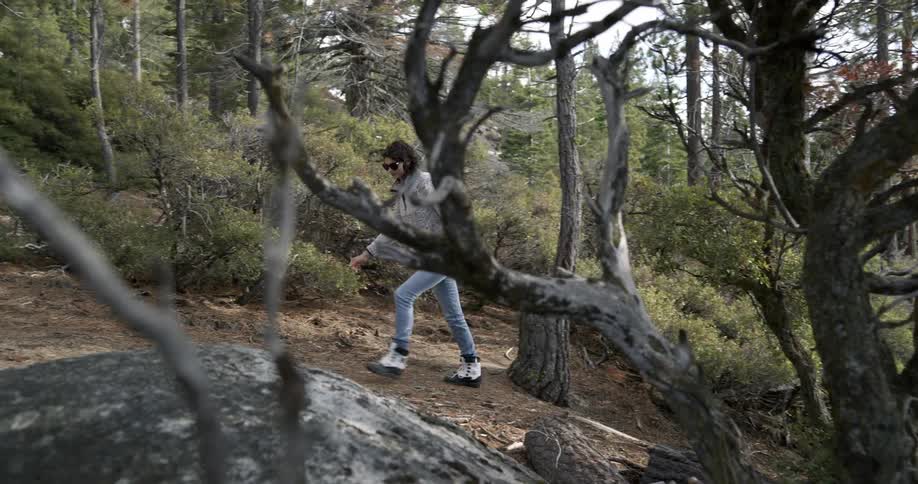 Woman with grey sweater and jeans and sunglasses walks uphill in the forest