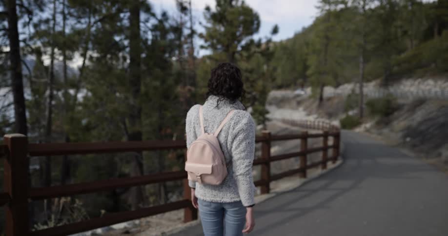 Woman with sunglasses a grey sweater a pink backpack and jeans walks down a cliff side road