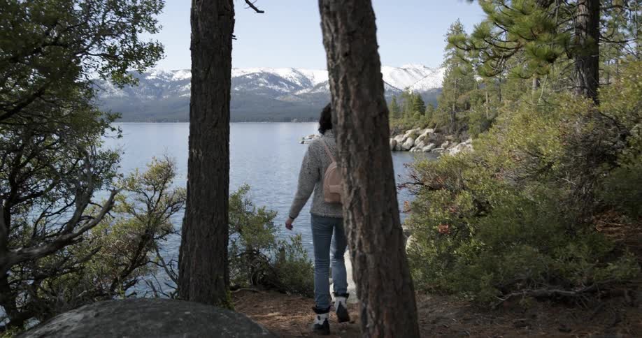 A woman with a backpack and sunglasses by a lake with a mountain range the sky and clouds in the background