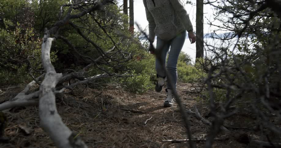 A woman wears white snow boots as she hikes down a trail in a forest near a lake