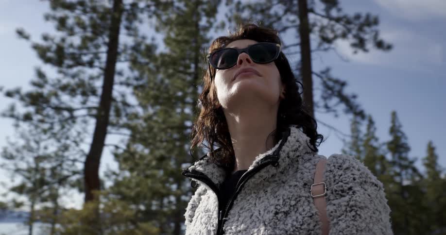A woman wearing large sunglasses looks up at the sky and trees as she stands between a lake and a slope