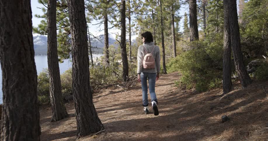 A woman wearing a bag on her back walks on a dirt trail looking at the lake