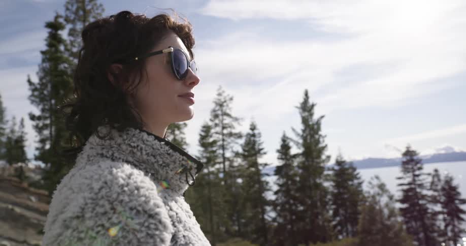 A woman with sunglasses and a jacket standing near a lake with trees the sky and clouds in the background