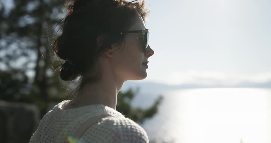 Woman stands under pine tree looking over ocean toward mountains and clouds on opposite shore