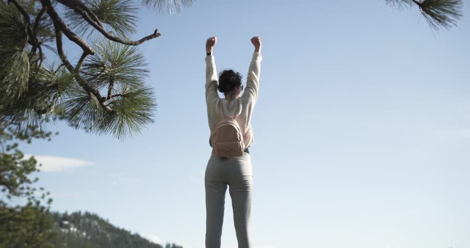 A woman climbing onto a boulder and raising her arms into the air while hiking in mountains