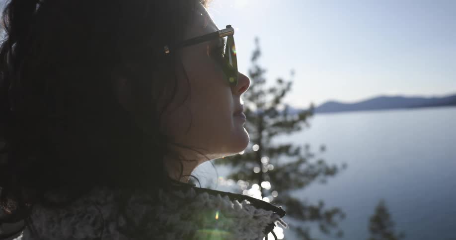 A woman sits on a park bench overlooking the water and trees below as the sun shimmers on the surface