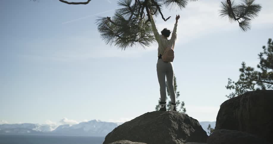 Young woman hiking in the mountains stands atop a rock in the summit celebrating