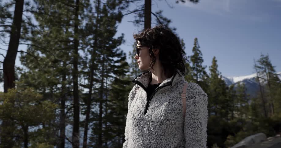 A woman with sunglasses and a jacket standing near a lake with trees the sky and clouds in the background