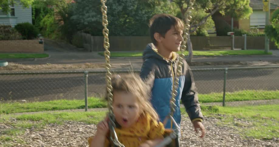 Boy pushes sister on swing in playground in neighborhood as grandmother films with mobile phone